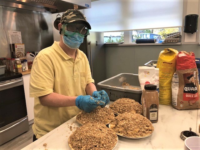 Person wearing a mask, gloves, and cap preparing multiple dishes topped with oats and crumble in a kitchen.