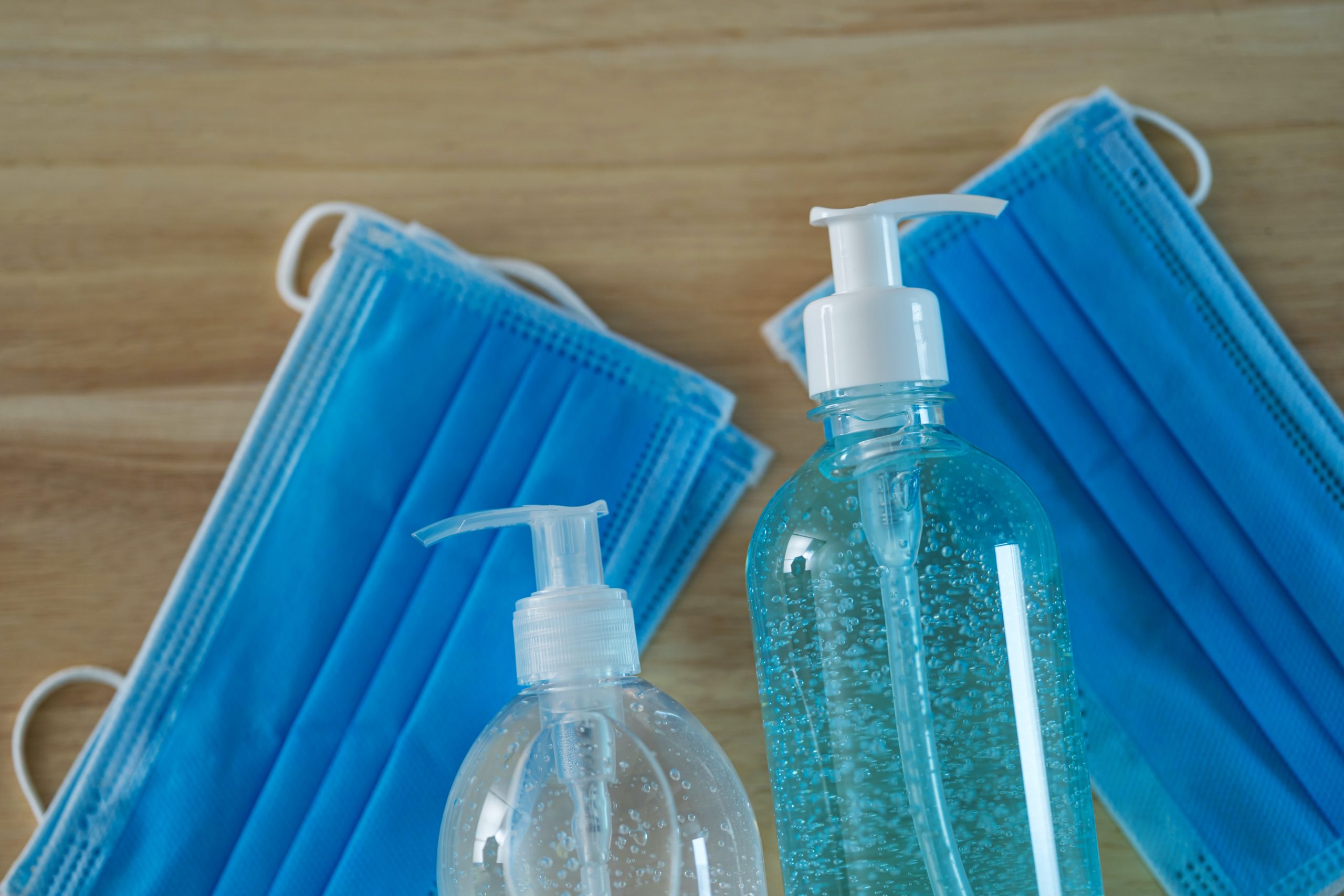 Two bottles of hand sanitizer placed beside blue disposable face masks on a wooden surface.