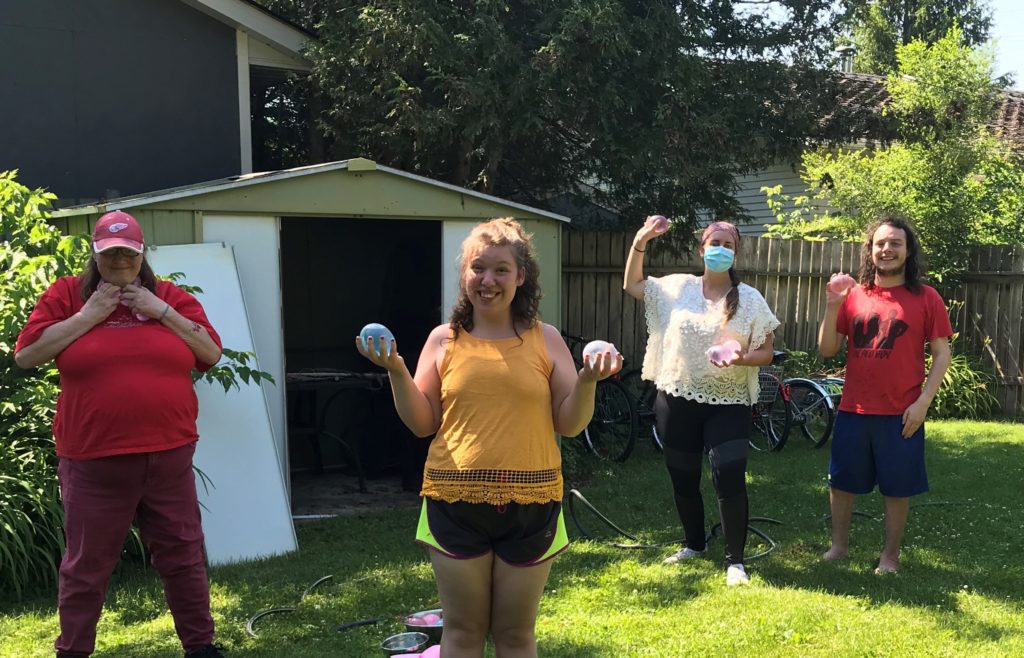 Four people outdoors smiling and holding water balloons during a summer activity at E3 Community Services.