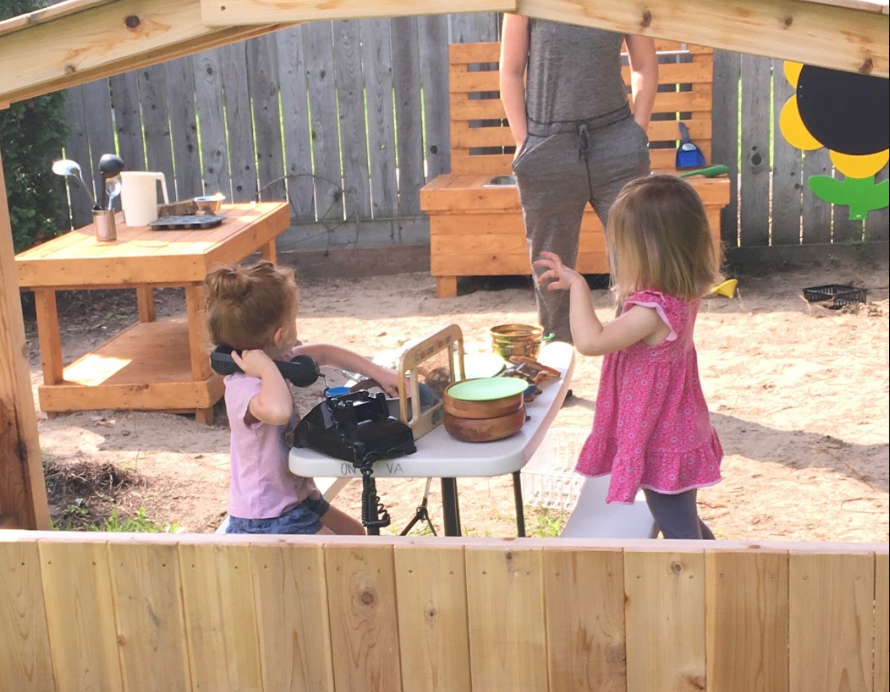 Two young girls playing in an outdoor mud kitchen setup. One child is holding a toy phone while the other gestures with her hand, with wooden play tables and utensils around them.