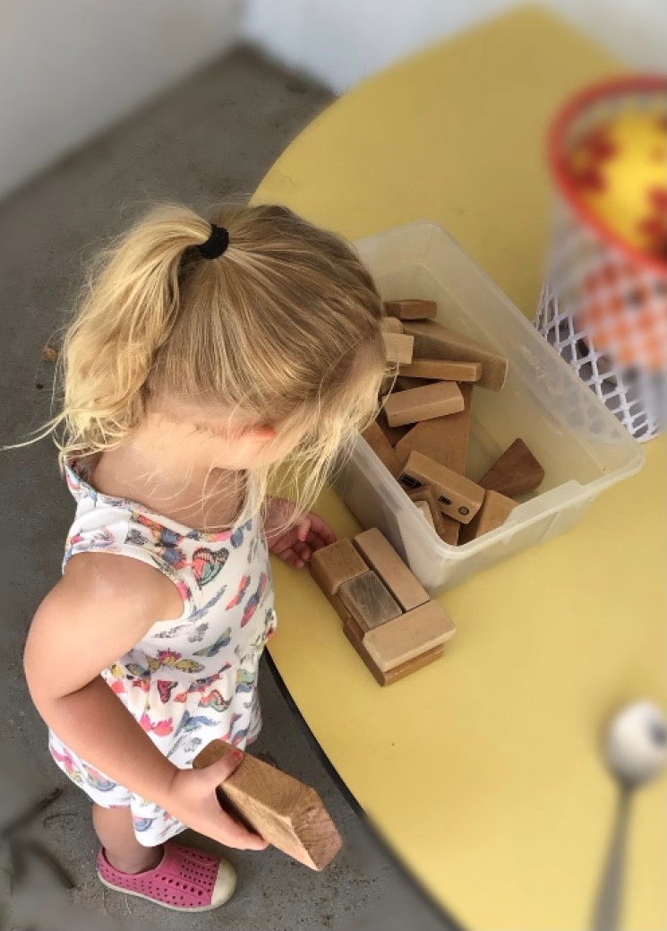 Young child playing with wooden building blocks on a yellow table, exploring pieces from a clear plastic bin.