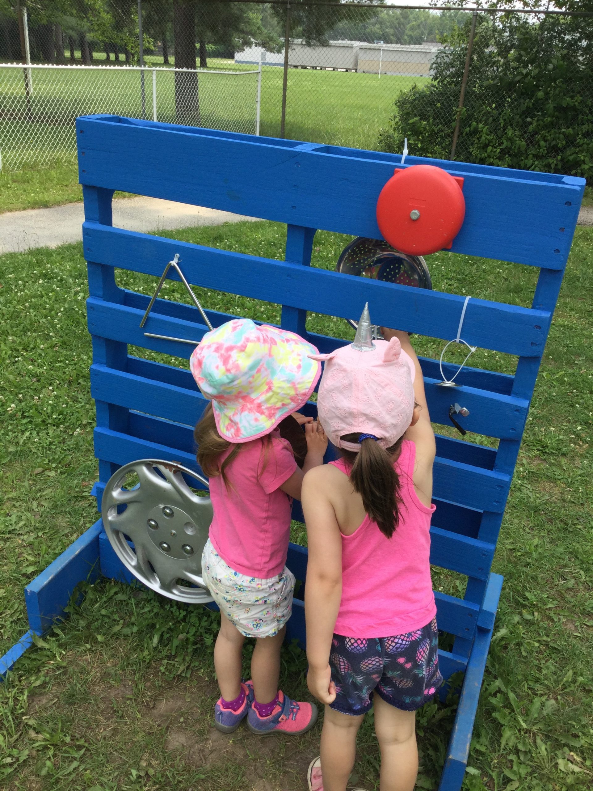 Two young girls in pink shirts and hats playing with a blue outdoor music wall made from a wooden pallet, featuring pots, pans, and other objects to make sounds.