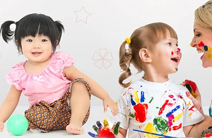 "Two young children and a caregiver engaging in playful activities, one child holding a ball and another covered in colorful hand paint during an art activity.