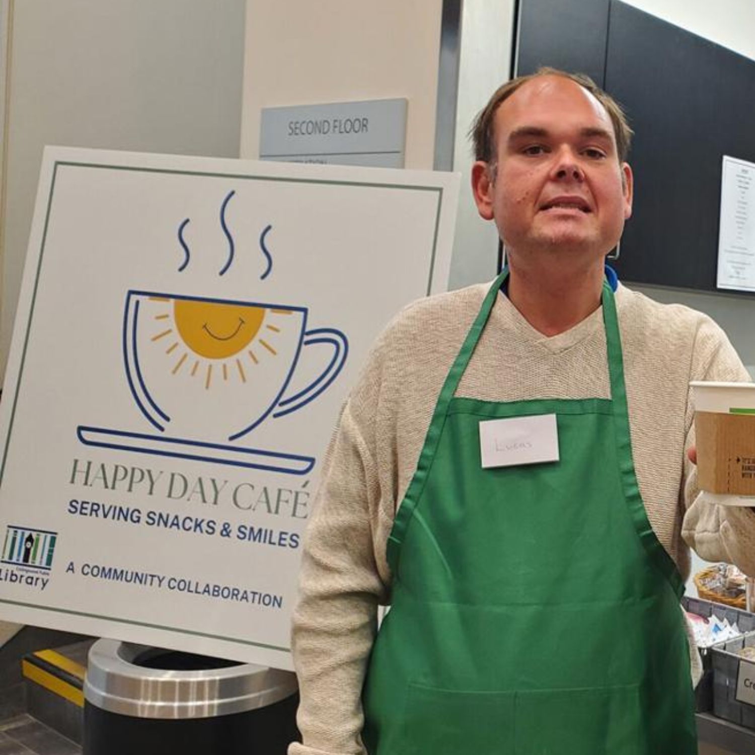 Man wearing a green apron and name tag serving coffee at Happy Day Café, with a sign in the background that reads 'Serving Snacks & Smiles – A Community Collaboration.'