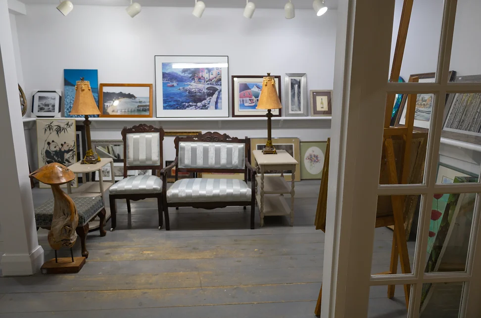 Cozy art gallery room with framed paintings on the walls and floor, vintage wooden chairs with striped cushions, two lamps on side tables, and rustic wooden floors.